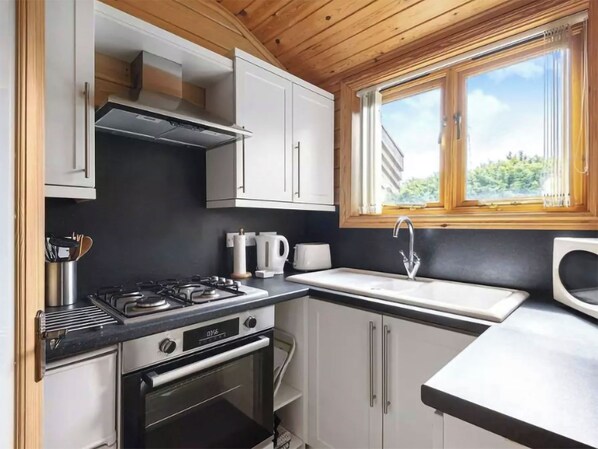 A compact kitchen with a gas stove, oven, white cabinets, and a window with a view of greenery.