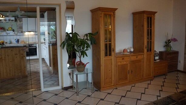 A spacious living area with a wooden cabinet, a glass table with a potted plant, and a kitchen in the background. - Gemütliche Erdgeschosswohnung mit Terrasse in Ortsrandlage in Waibstadt (Waibstadt)