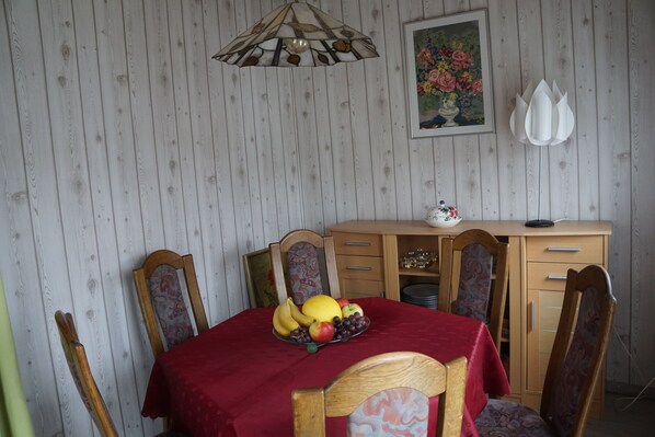 A dining area with a wooden table covered by a red tablecloth, surrounded by wooden chairs with patterned cushions. A fruit bowl with bananas, apples, and grapes is on the table. - Gemütliche Erdgeschosswohnung mit Terrasse in Ortsrandlage in Waibstadt (Waibstadt)