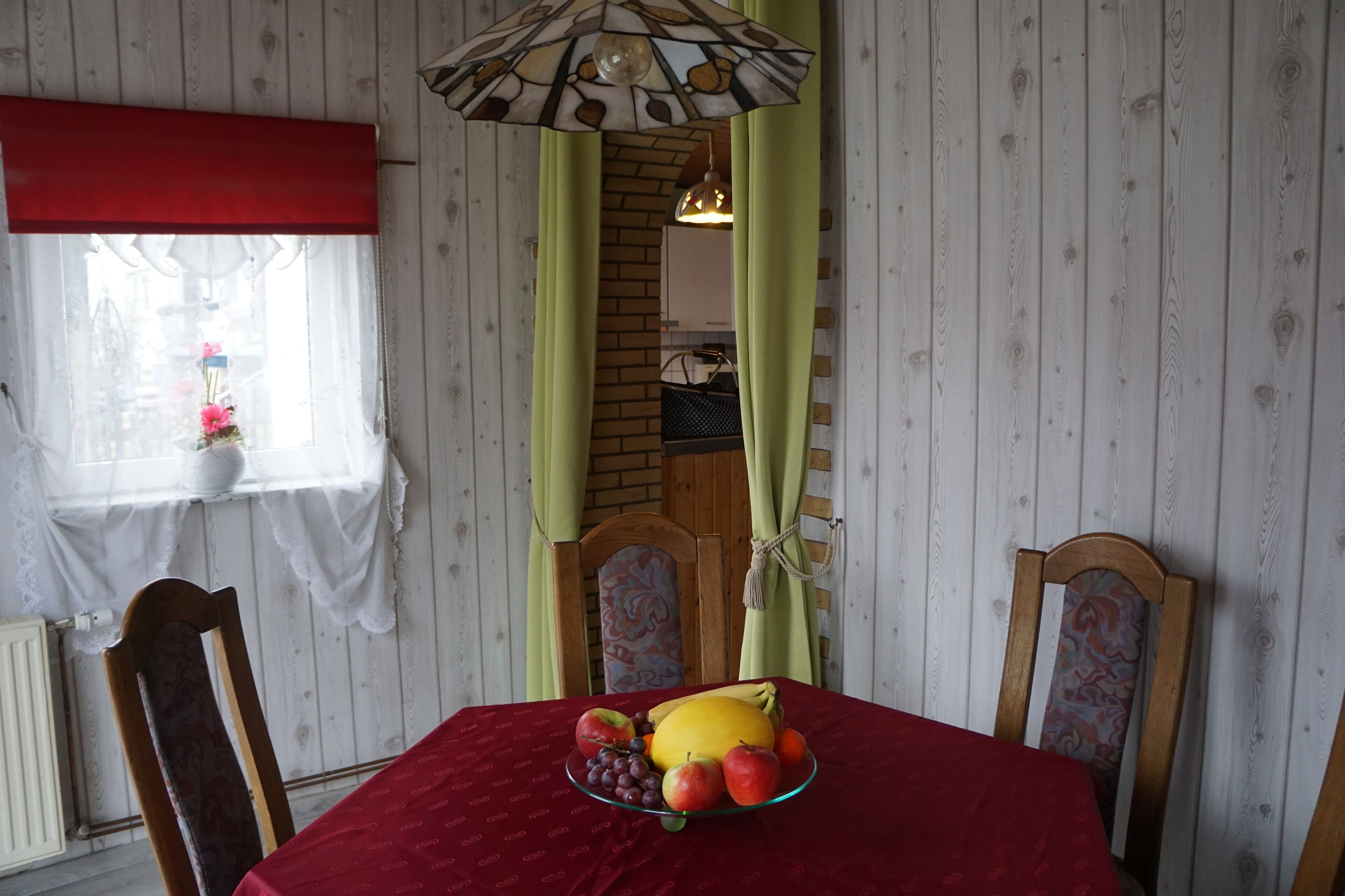 A dining table with a red tablecloth, surrounded by wooden chairs with patterned upholstery. A fruit bowl with apples, grapes, and a melon is placed on the table.