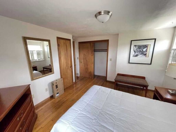 Quarto com cama, guarda-roupa de madeira, um banquinho, um quadro emoldurado e um espelho.