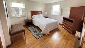 Un dormitorio con suelo de madera, una cama con ropa blanca, una mesita de noche, un aparador y dos ventanas con contraventanas.