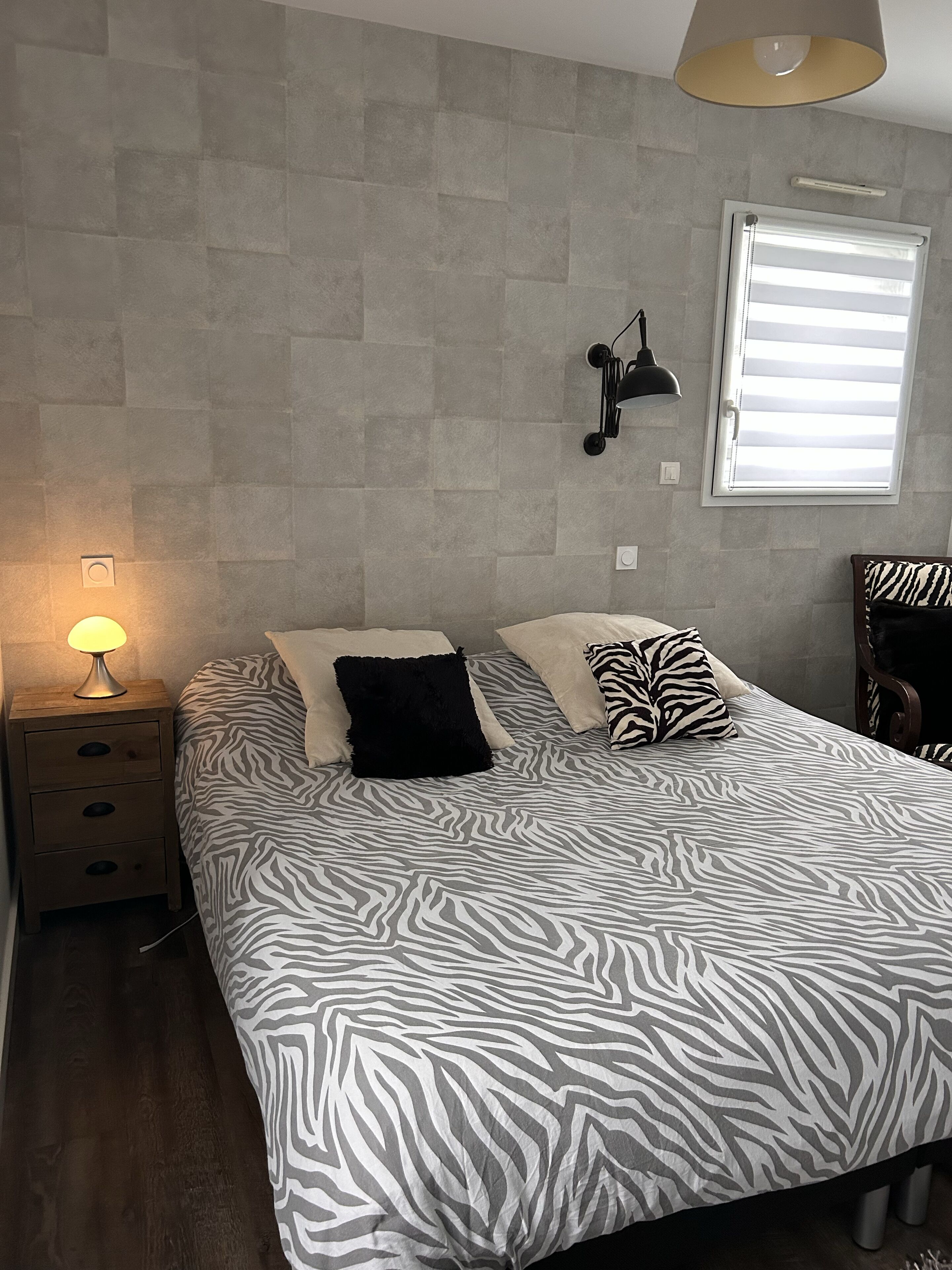 Een slaapkamer met een bedovertrek met zebraprint, een bedlamp, een wandlamp en een raam met jaloezieën.