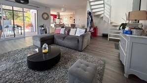 A modern living room with a grey sectional sofa, a black coffee table, and a grey ottoman. There is a staircase leading to an upper level, and a dining area with a red wall and a kitchen in the background. - Maison Moderne Avec Piscine Chauffée, à 1km de L'avenue de Gaulle à La Baule (Guérande)