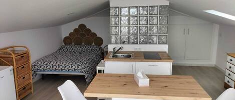A compact, modern loft bedroom with a sloped ceiling, a bed with a patterned bedspread, a kitchenette with a sink and cabinets, and a dining area with a wooden table and two white chairs.