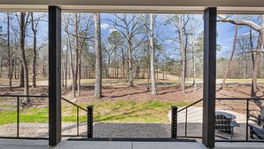 A view from a covered outdoor area with a metal railing, looking out onto a forested park area with trees and a barbecue grill. - Newly built Chalet on State Park Golf Course 5 min from Pickwick Lake! (Counce)