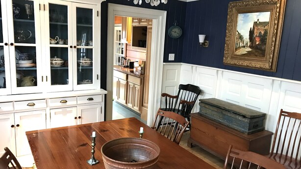 Une salle à manger avec une table en bois, des chaises, un grand saladier et un buffet aux portes vitrées.
