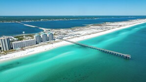 Vista aérea de una zona costera con un muelle largo que se extiende hacia aguas turquesas, rodeada por una playa y varios edificios altos.
