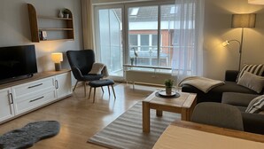 A modern living room with a flat-screen TV, a dark armchair, a sofa, a wooden coffee table, and a large window with a view of buildings. - Gemütliche Ferienwohnung Mitten im Zentrum von Büsum (Büsum)