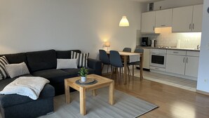 A modern living room with a dark sofa, a small wooden table with a plant, and a dining area with chairs and a table. A kitchen with white cabinets and a stove is visible in the background. - Gemütliche Ferienwohnung Mitten im Zentrum von Büsum (Büsum)