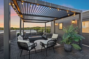 Een overdekt terras met een bank, stoelen en een tafel, omgeven door een pergola met lichtslingers.