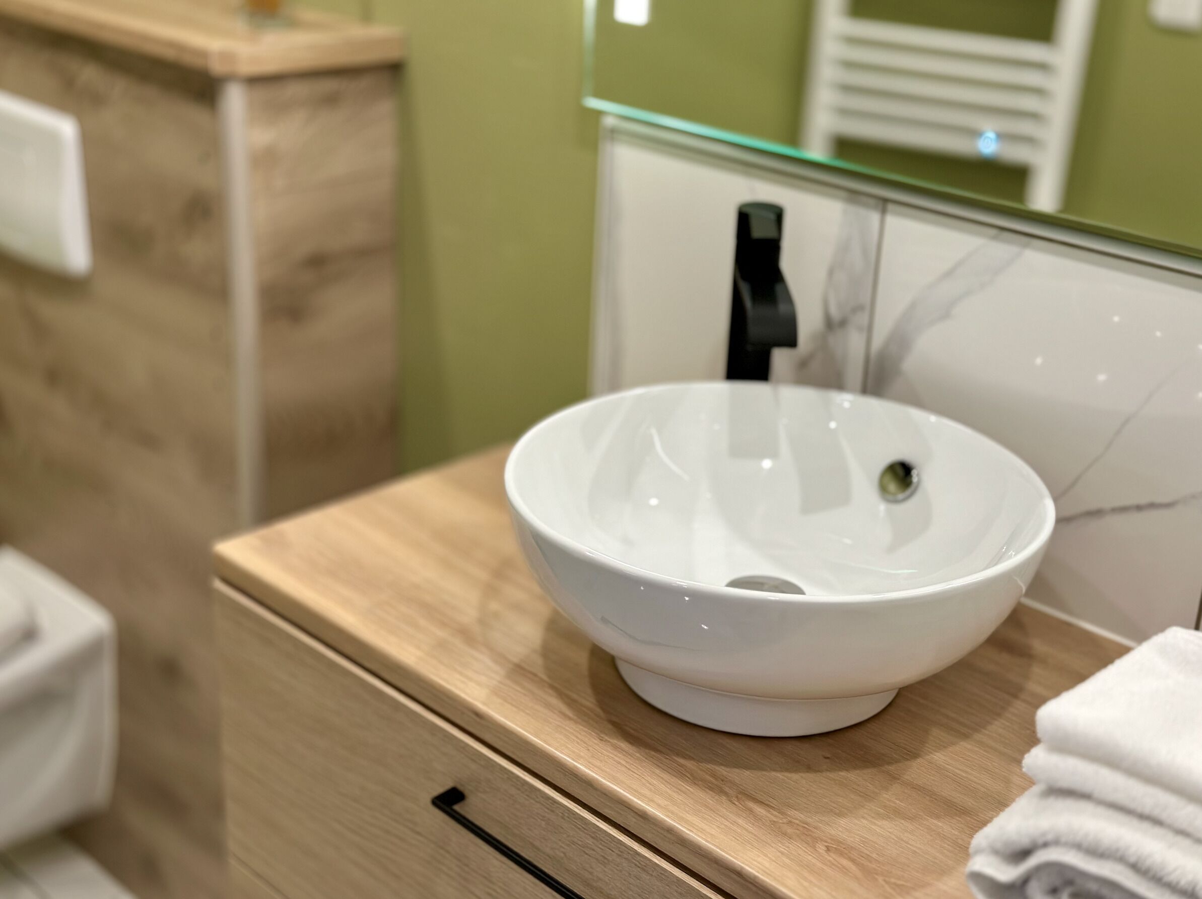 Une salle de bain avec un lavabo sur colonne blanc, un meuble-lavabo en bois, un porte-serviettes et un miroir devant un mur vert.