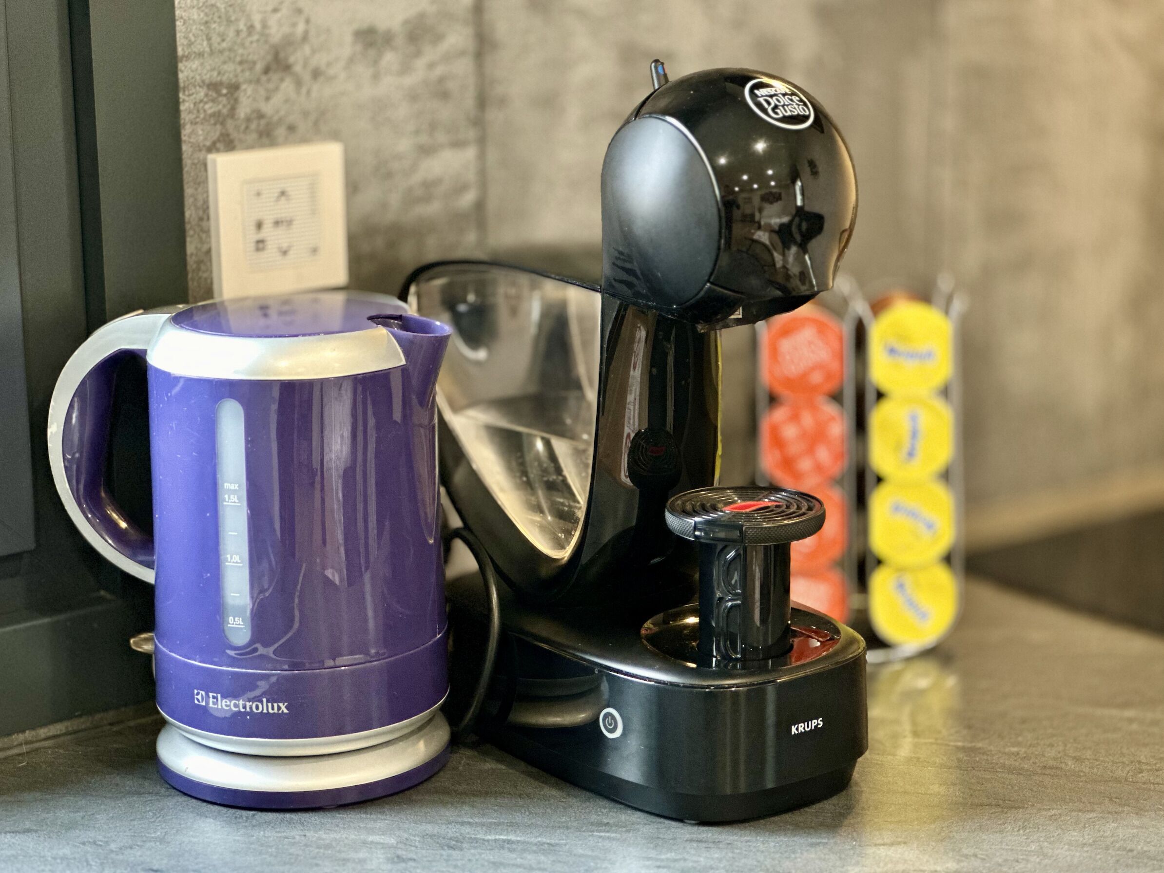 En kaffemaskine, en lilla kop og et sæt gule kopper på en bordplade.