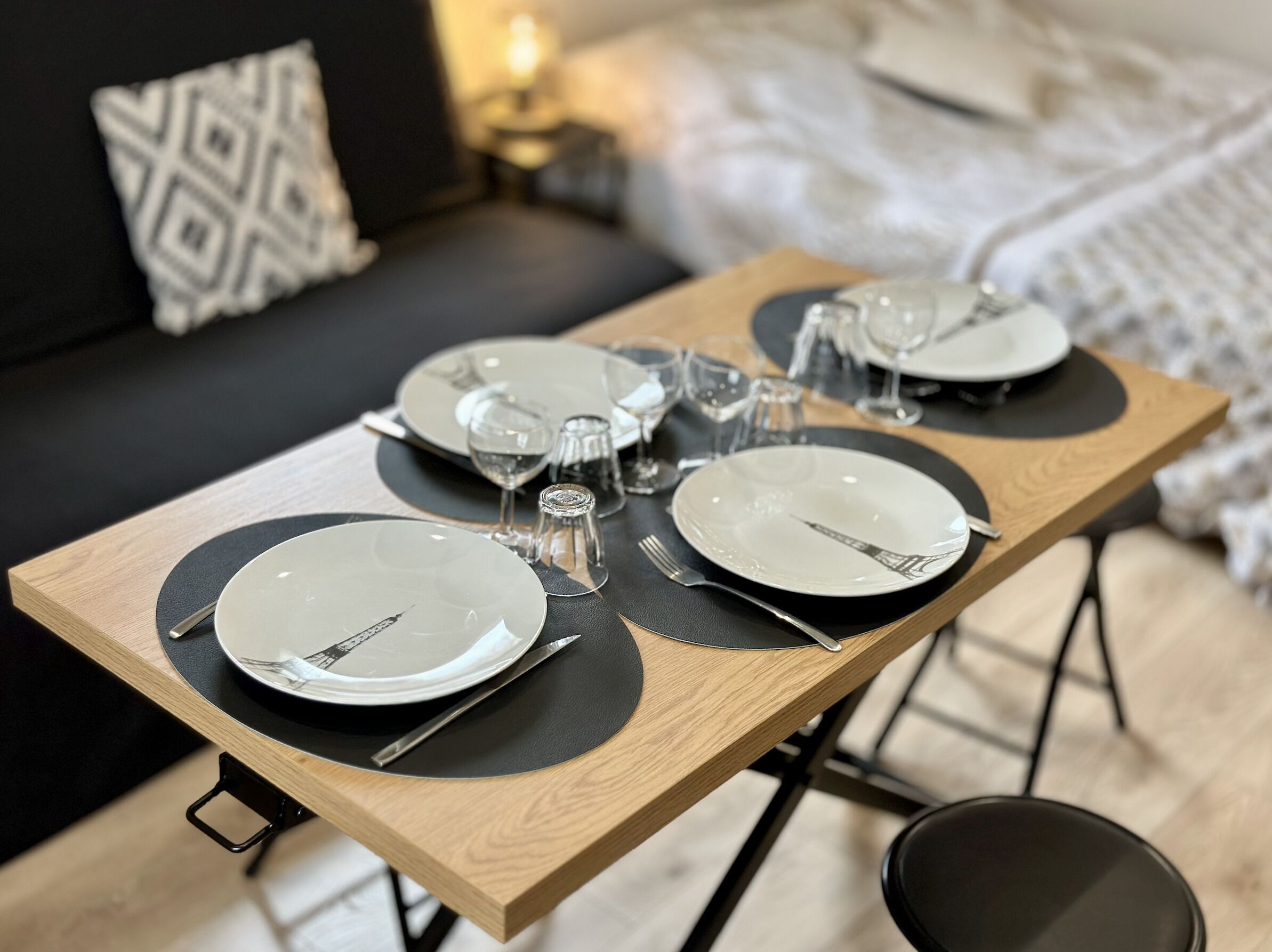 Une table à manger élégamment dressée, avec des assiettes, des couverts et des verres, placée dans une pièce moderne où l’on aperçoit un canapé et une lampe en arrière-plan.
