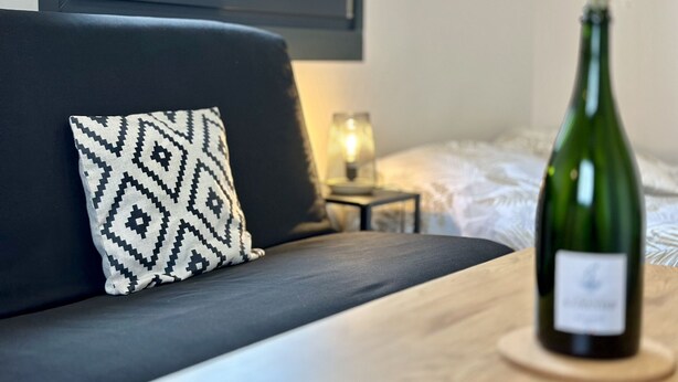 A modern room with a dark sofa, a patterned pillow, a bedside table with a bottle, and a window with a view.