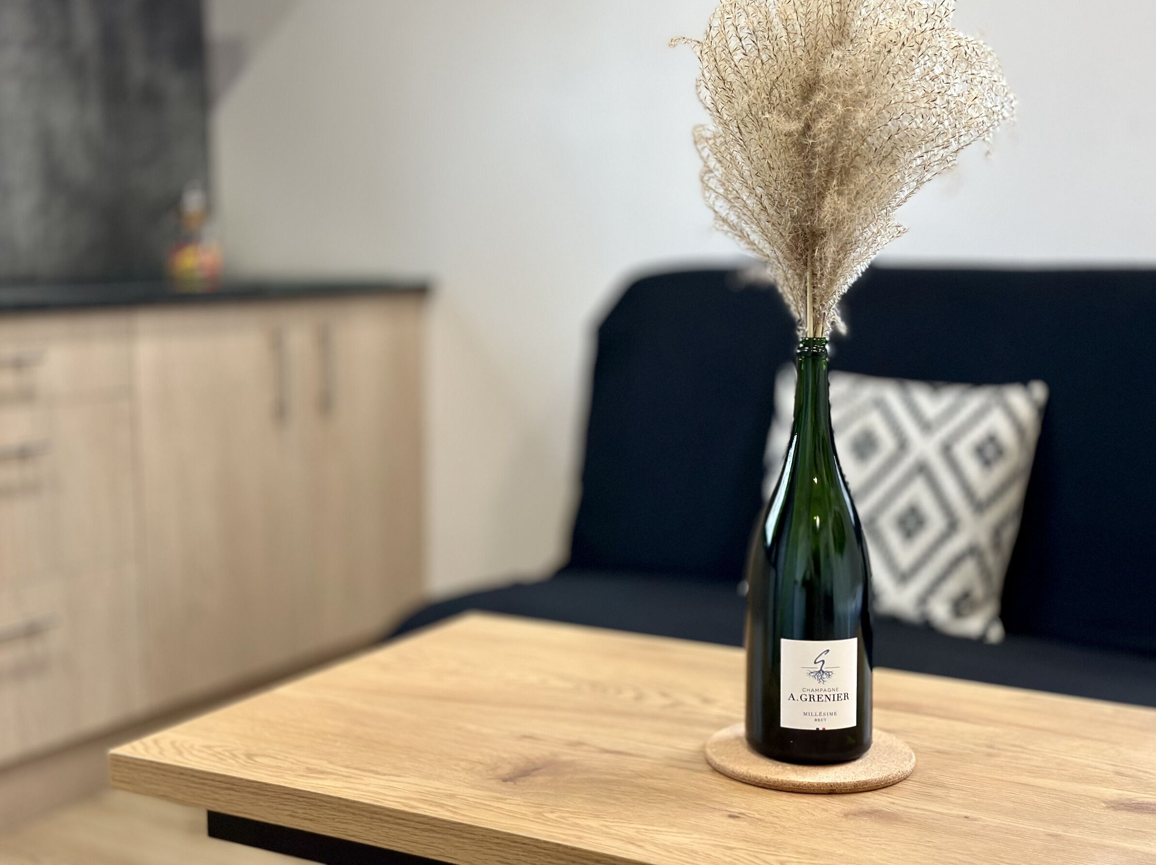 A bottle of A. Grenier champagne on a wooden table with a dark blue sofa and a patterned pillow in the background.