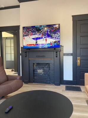 Una sala de estar con chimenea, un televisor sobre ella mostrando un evento deportivo, un sillón de cuero marrón y un reposapiés negro.