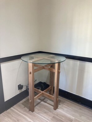 Una mesa en esquina con superficie de vidrio y estructura de madera, ubicada junto a una pared con zócalo negro.