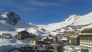 Un pueblo nevado a los pies de una montaña, con edificios, un estacionamiento y un cielo azul despejado.