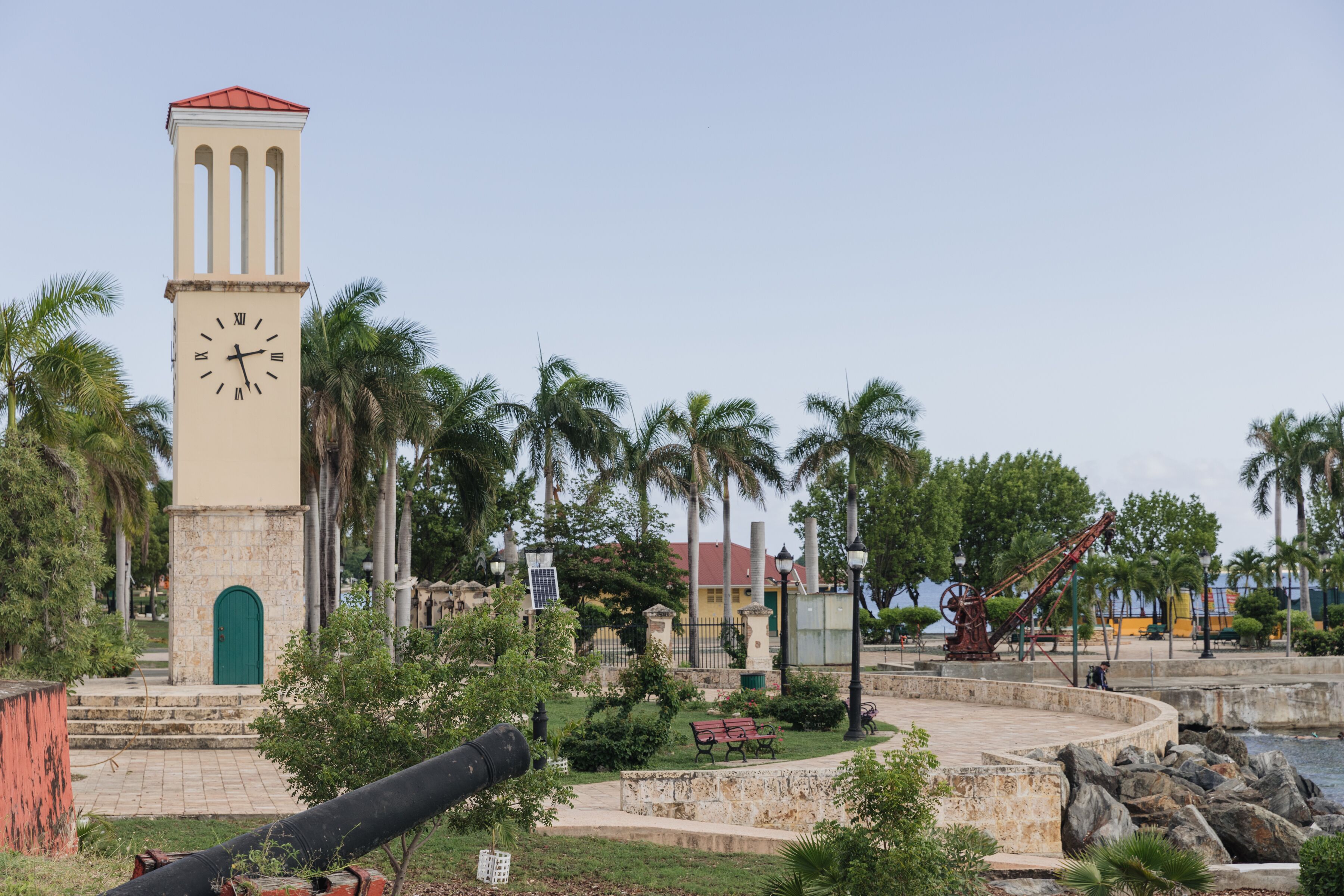 Une tour de l’horloge avec une porte verte, entourée de palmiers et d’un parc agrémenté de bancs et d’une fontaine.