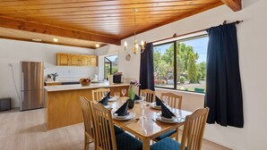 Un comedor con una mesa de madera dispuesta para cuatro personas, rodeada de sillas con cojines azules. Se ve una cocina con armarios de madera y un refrigerador de acero inoxidable al fondo.