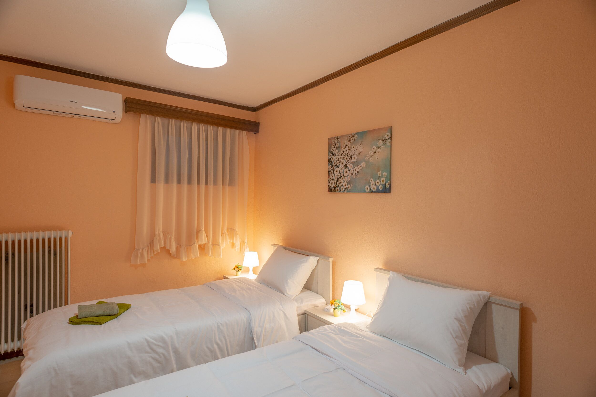 Hotellihuone, jossa on kaksi sänkyä, patteri, ilmastointilaite, ikkuna verhoilla ja taulu seinällä.