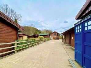 A row of wooden cabins with a concrete path, a blue police box, and a wooden fence. - Pine Lodge - UK46279 (Camerton, near Bath)