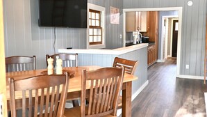 A dining area with a wooden table and chairs, a TV mounted on the wall, and a kitchen in the background. - Waterfront house on Miner Lake- Waupaca Chain (Waupaca)