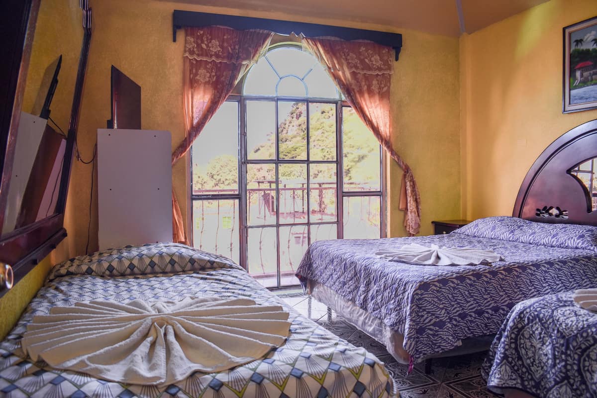 A room with two beds, a wooden headboard, a window with curtains, and a wall-mounted TV.