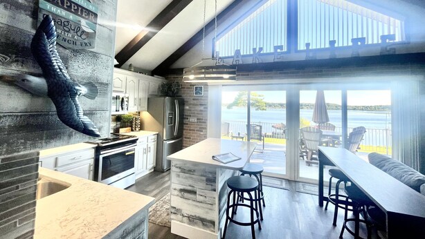 A modern kitchen with a large island, a dining area with bar stools, and a view of the lake through large sliding doors. - Lincoln Lake Paradise with Tiki Bar & Hot Tub (Gowen)