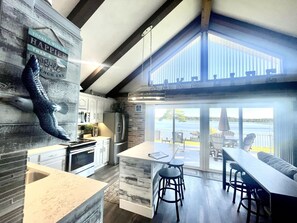 Une cuisine moderne dotée d’un grand îlot central, d’un coin repas avec des tabourets de bar et offrant une vue sur le lac grâce à de grandes portes coulissantes.
