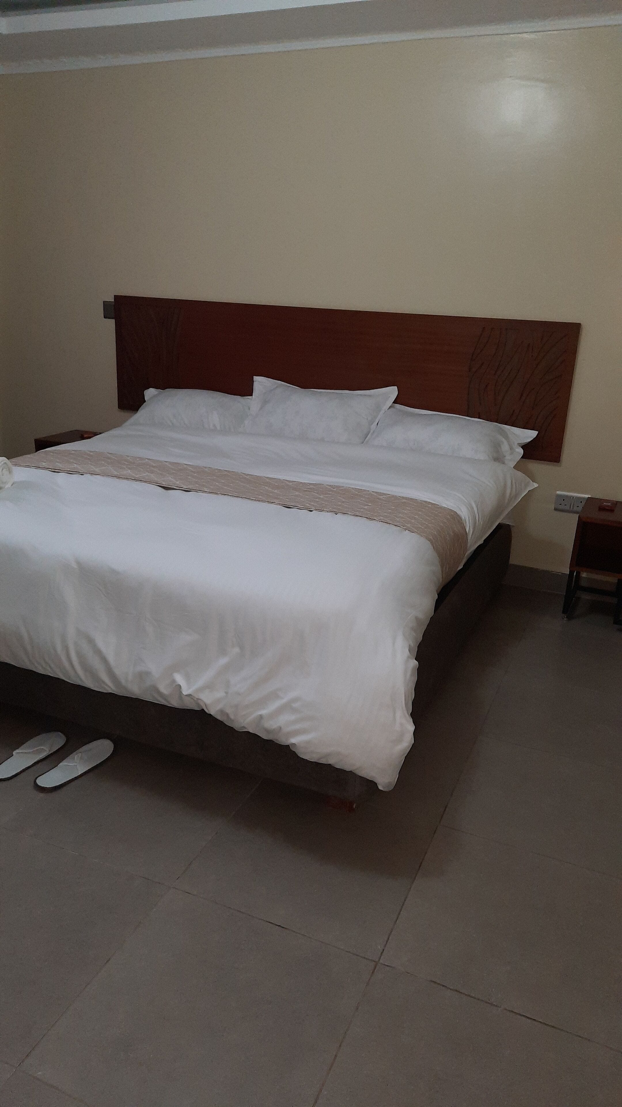 A neatly made bed with white linens and a wooden headboard, a small bedside table with a lamp, and a pair of slippers on the floor.