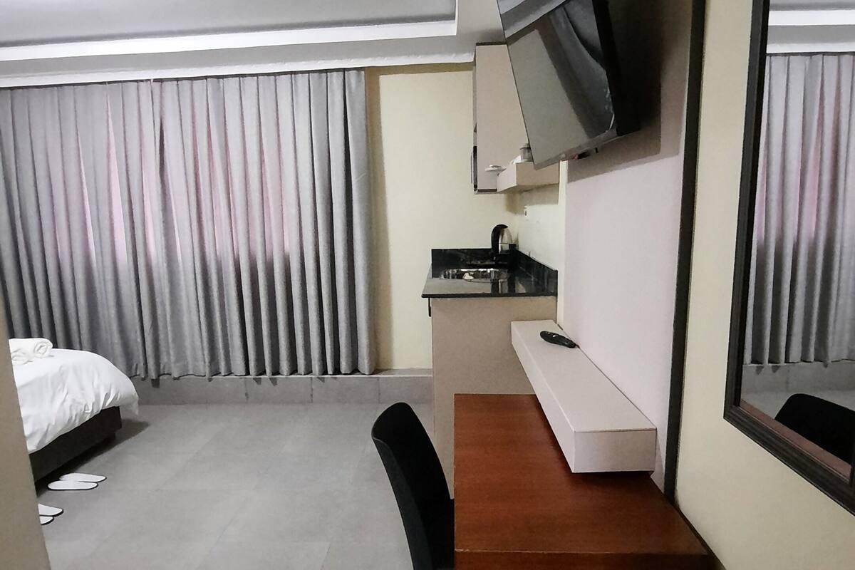 Una habitación de hotel con cama, escritorio, silla, televisor montado en la pared y ventana con cortinas.