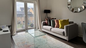 Un salón moderno con una mesa de centro de vidrio, un sofá con cojines de colores, un espejo grande en la pared y vistas al exterior a través de una puerta corredera.