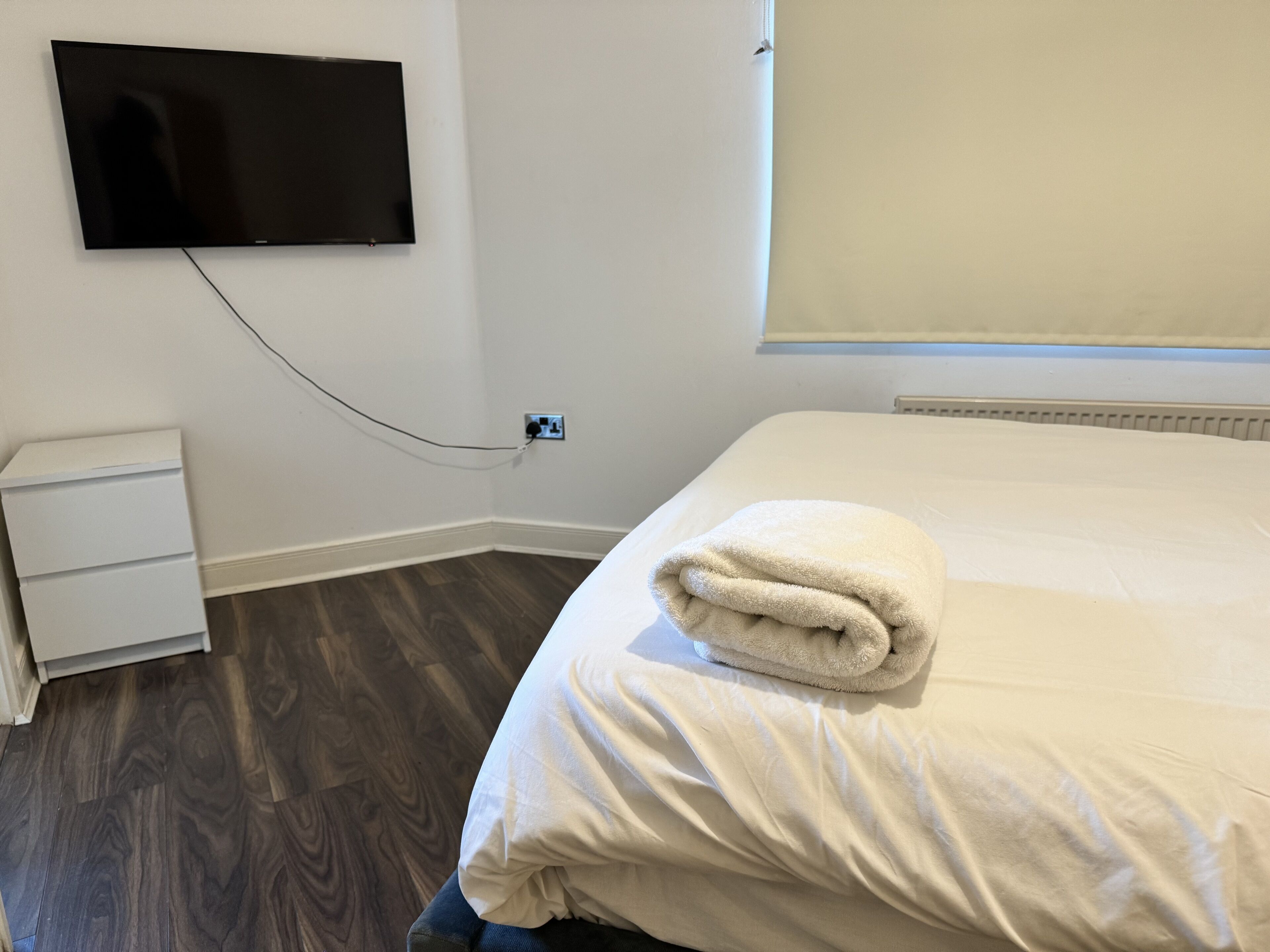 Une chambre d’hôtel équipée d’un téléviseur à écran plat fixé au mur, d’une table de chevet, d’un lit soigneusement fait avec une serviette pliée, et d’une fenêtre avec des stores.