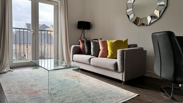 Una sala moderna con un sofá, cojines coloridos, una mesa de centro de vidrio, un espejo grande y vistas a través de una puerta corrediza de cristal.