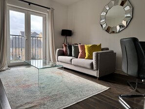 A modern living room with a sofa, colorful cushions, a glass coffee table, a large mirror, and a view through a sliding glass door. - Havana 2 bed Apartment in london (London)