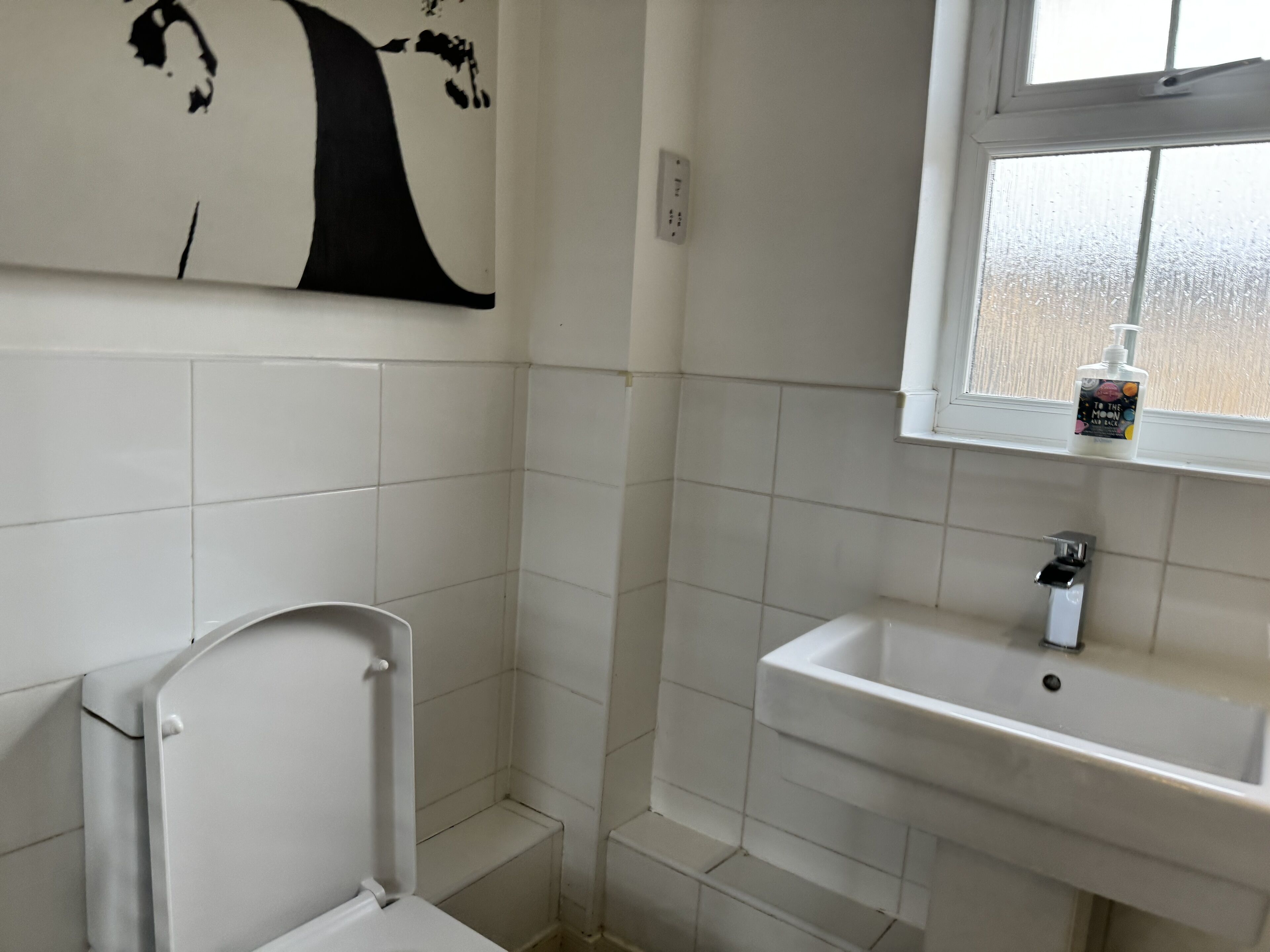 A bathroom with white tiles, a toilet, a sink, and a window with a bottle of hand sanitizer.