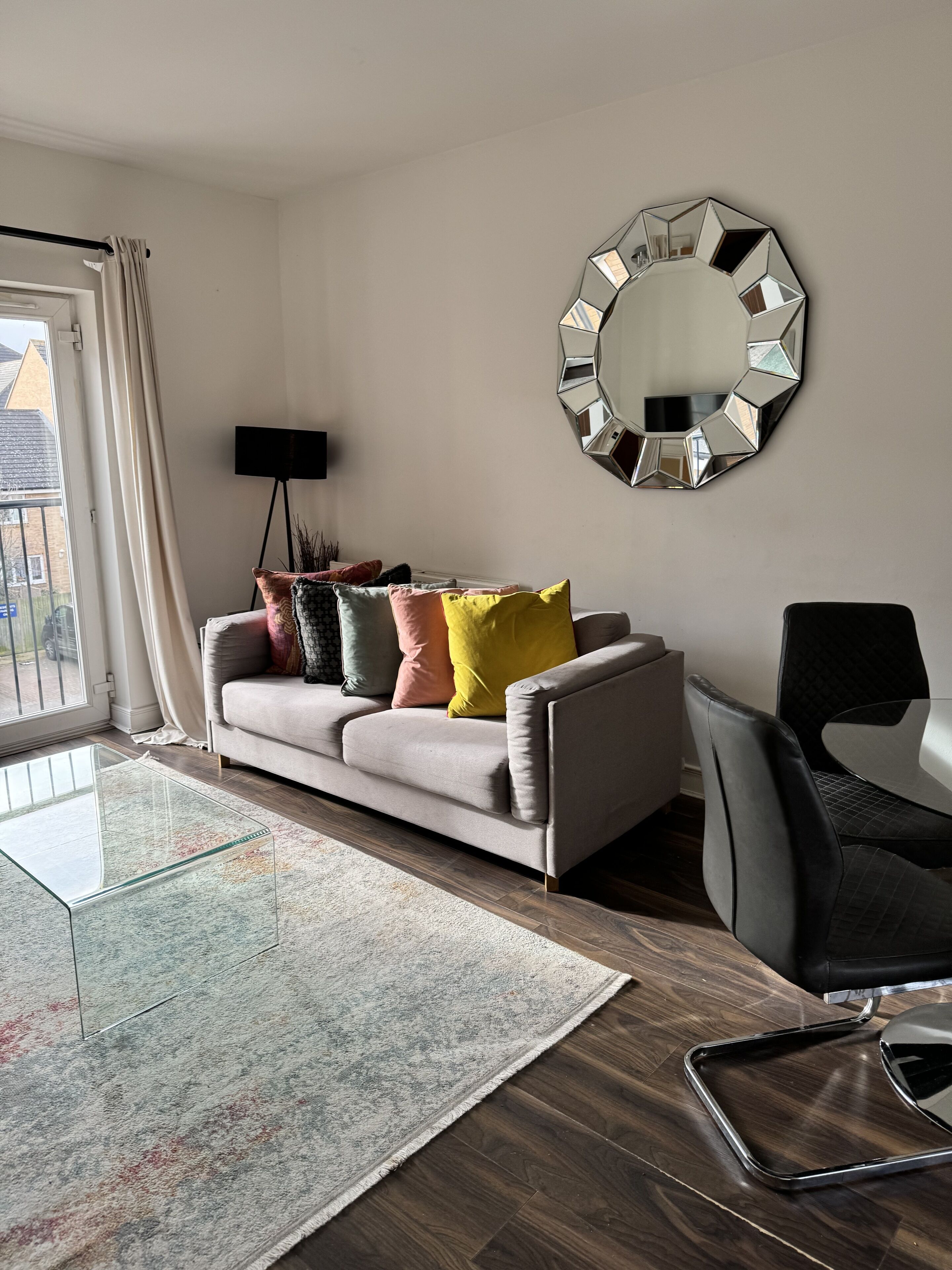 A living room with a sofa, colorful cushions, a round mirror, a black chair, and a glass coffee table.