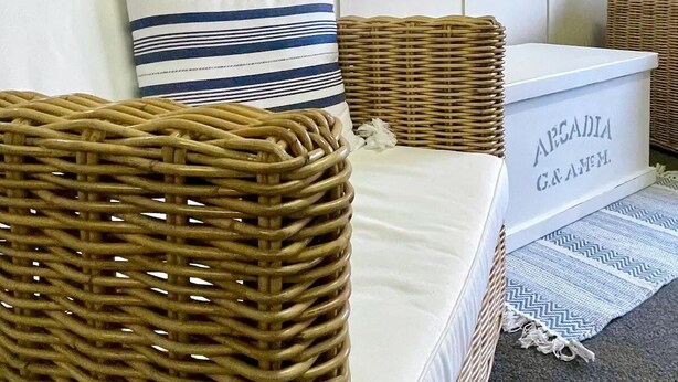 Un canapé en osier avec un coussin blanc et un oreiller à rayures, un panier en osier et un tableau encadré représentant une femme portant un chapeau blanc.