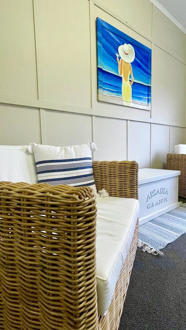 Un canapé en osier avec un coussin blanc et un oreiller à rayures, un panier en osier et un tableau encadré représentant une femme portant un chapeau blanc.