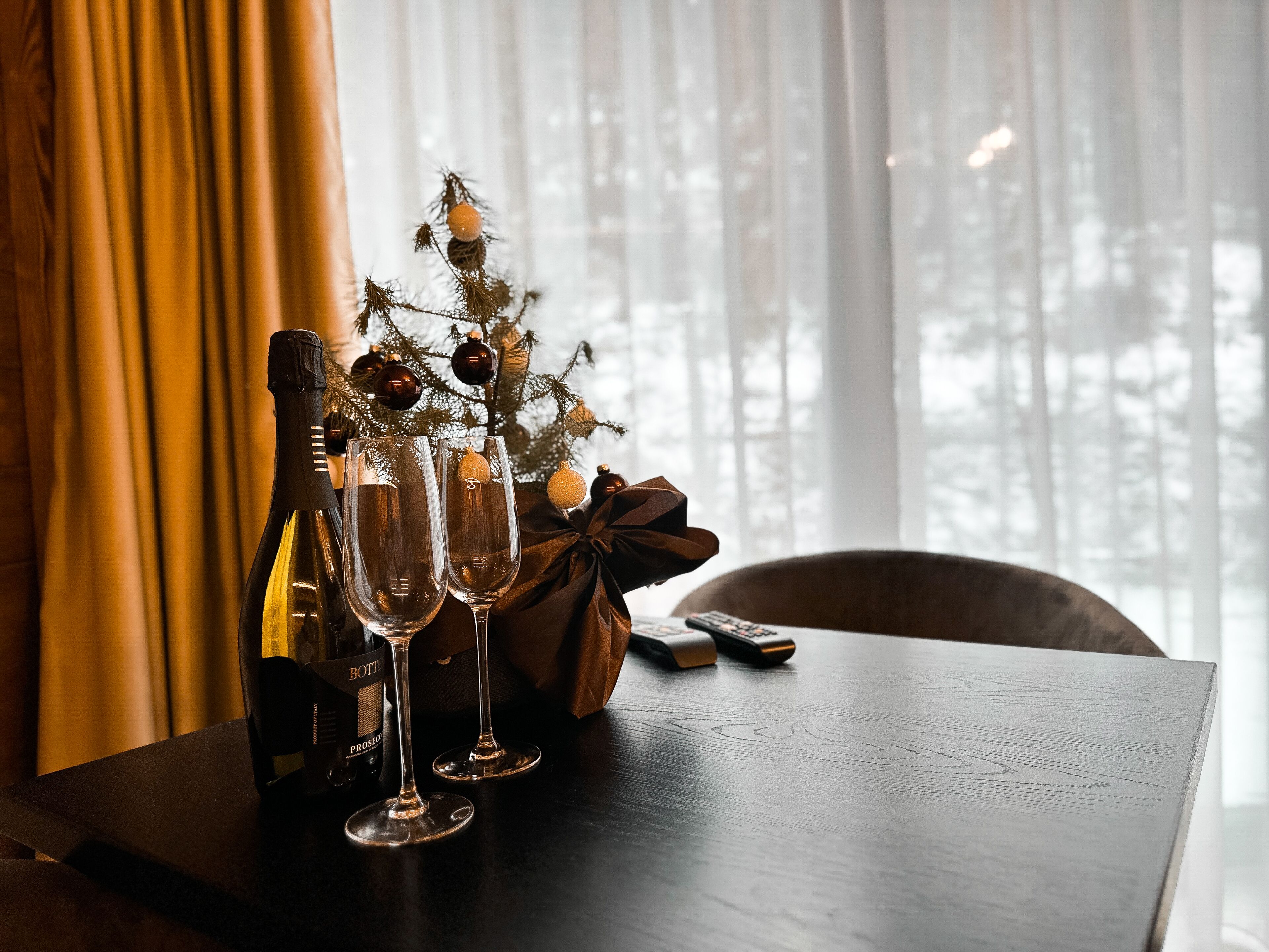 A table with a bottle of wine, two glasses, a small Christmas tree, and a gift box.