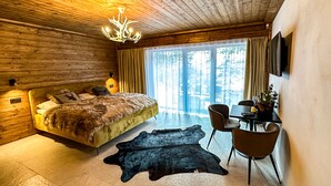A modern bedroom with a wooden ceiling, a large bed with a fur throw, a desk with a chair, and a view of trees through large windows. - Jasna Boutique Chalet (Demanovska Dolina)