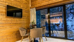 A modern interior with wooden paneling, a flat-screen TV mounted on the wall, a dining area with two chairs, and a large window offering a snowy forest view. - Jasna Boutique Chalet (Demanovska Dolina)