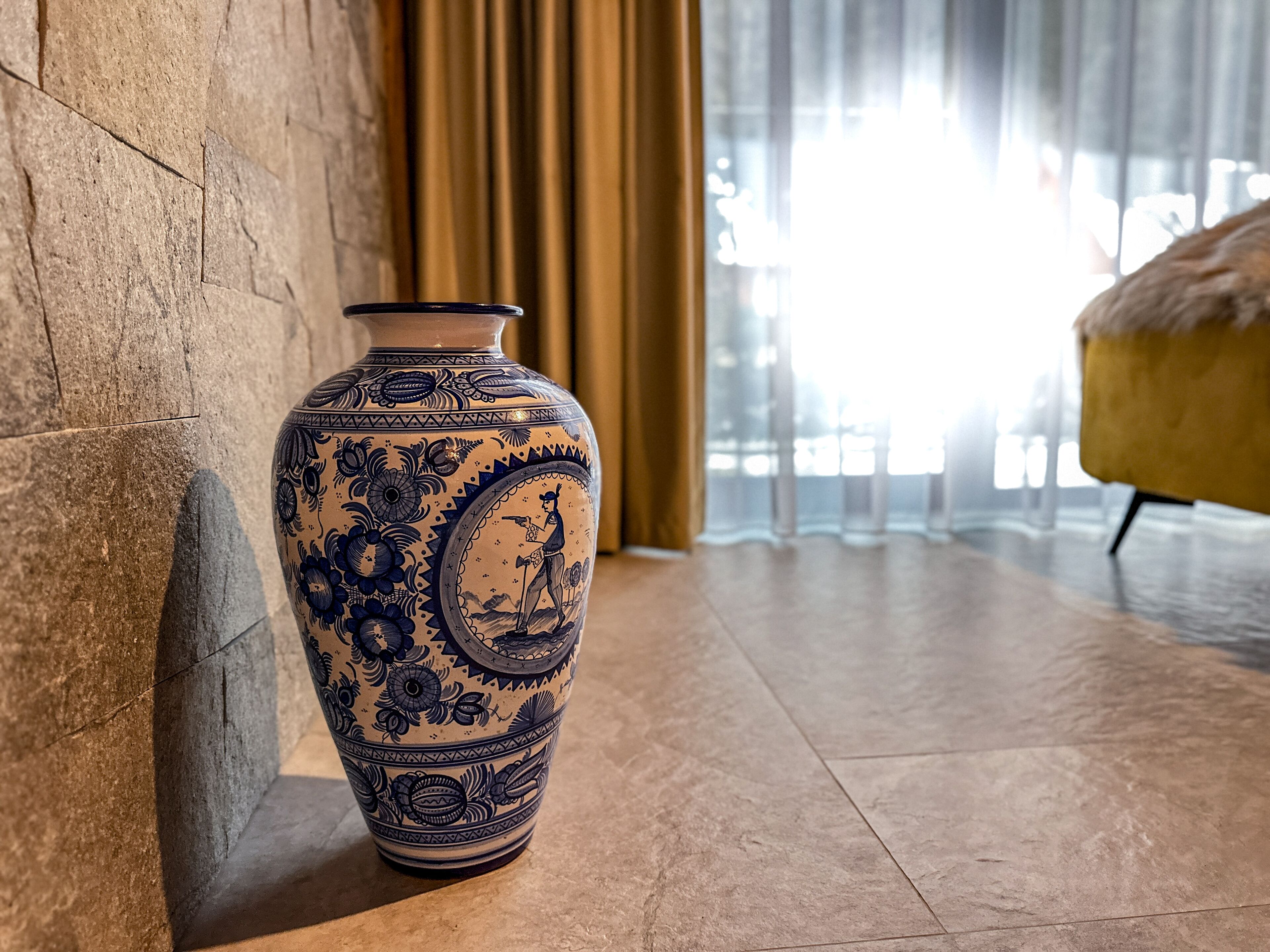 A large ceramic vase with intricate blue designs on a stone floor near a window with curtains.