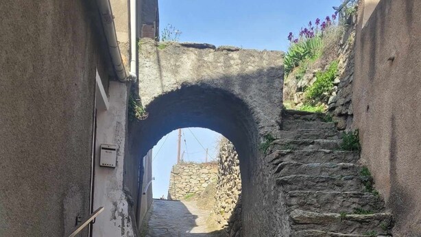 Un étroit sentier de pierre mène à travers une arche en pierre, bordé de murs et d’escaliers.