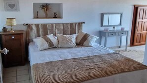 Une chambre avec un plafond en bois, un lit recouvert d’une couverture à motifs, une table de chevet avec une lampe, une commode et un miroir.