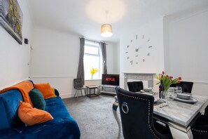 Ein modernes Wohnzimmer mit einem blauen Sofa, einem Esstisch gedeckt mit Tellern, Gläsern und einer Blumenvase. Ein Flachbildfernseher ist an der Wand befestigt und ein Kamin ist im Hintergrund zu sehen.