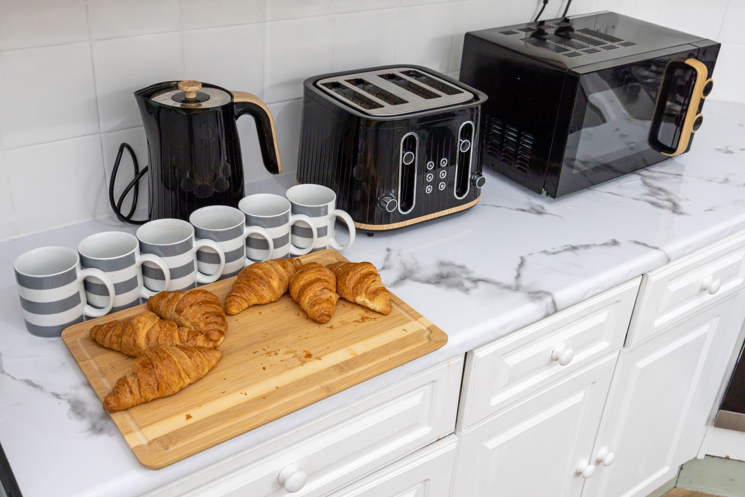 A kitchen counter with a toaster, a coffee maker, and a microwave. There are several mugs and croissants on the counter.