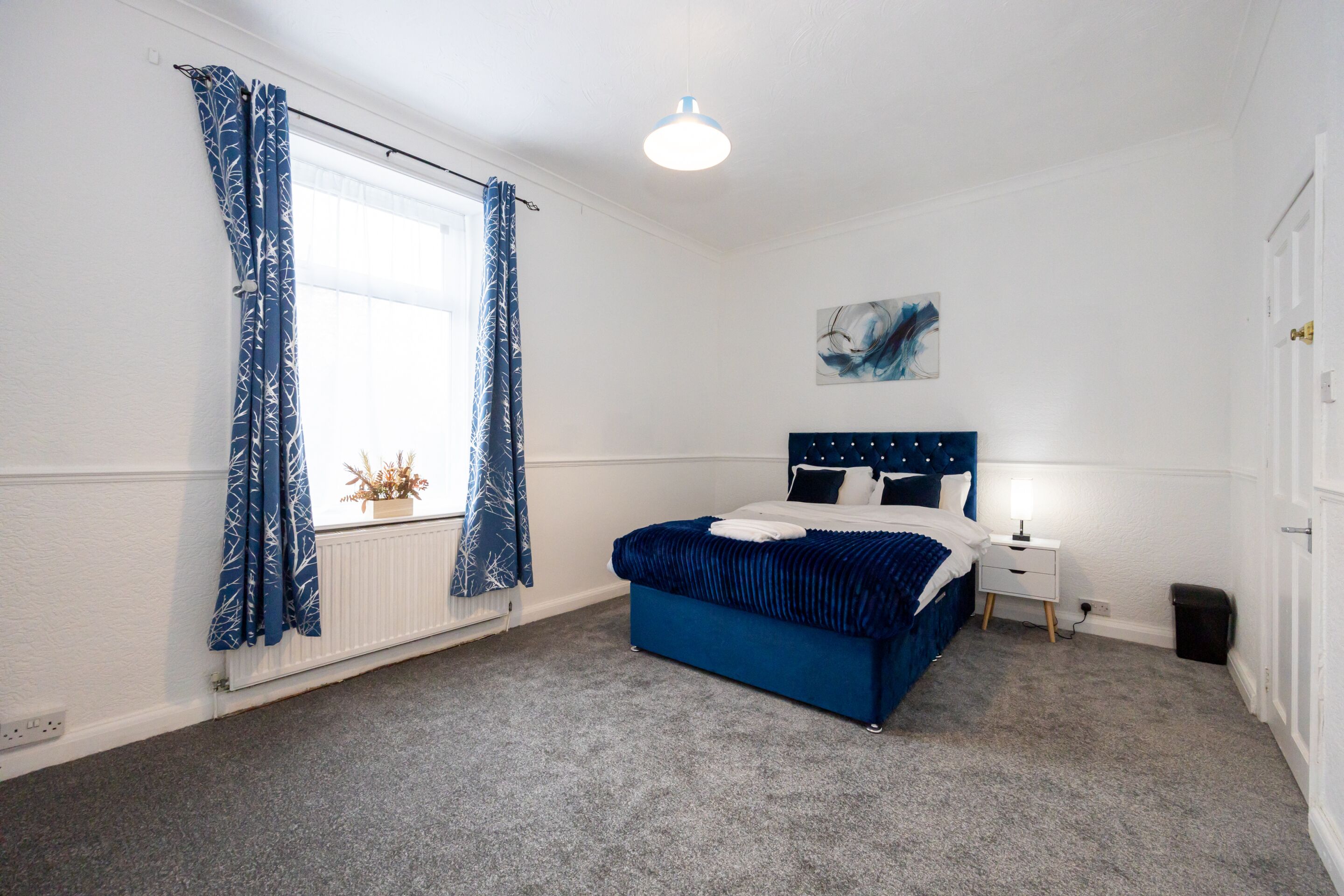 A bedroom with a bed, a bedside table, a lamp, a window with curtains, a radiator, and a wall-mounted artwork.